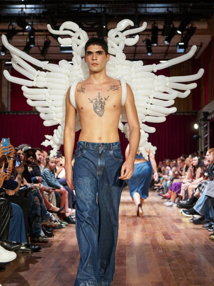 Homem de pele clara tatuado sem camisa, com calça jeans e bexiga que imita a logo da marca nas costas