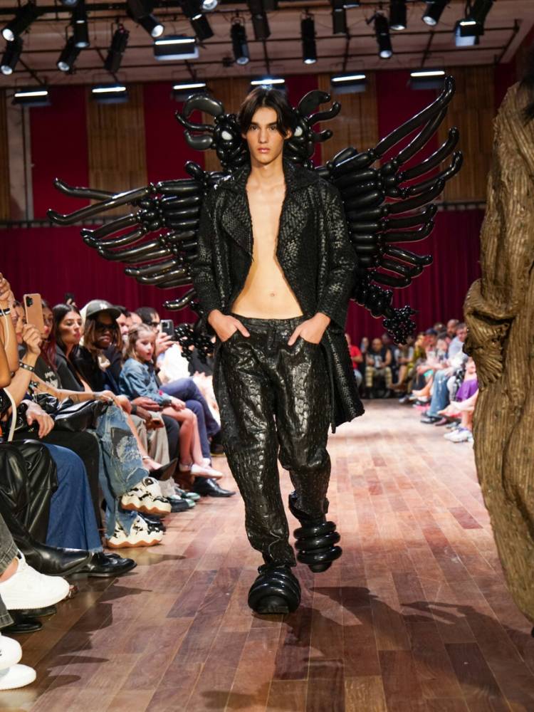 Homem de pele clara usando jaqueta e calça de couro pretas, além de logo da marca feito de bexiga nas costas
