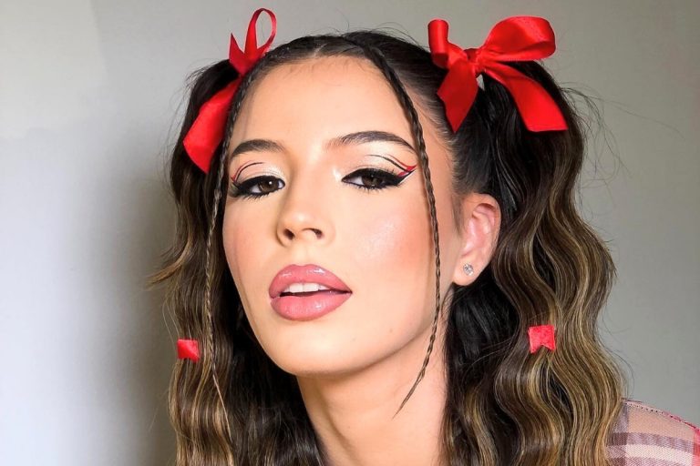 Uma jovem com maquiagem festiva, destacando um delineado gráfico preto com um toque de vermelho no canto externo. Ela tem lábios rosados e um penteado com duas marias-chiquinhas, adornadas com laços vermelhos vibrantes e pequenas fitas presas no cabelo.