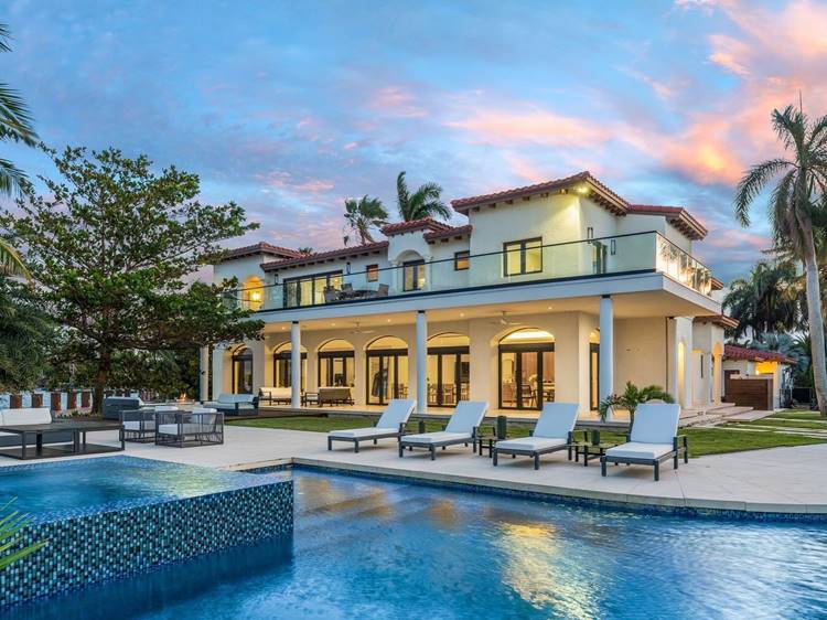 Uma mansão luxuosa com dois andares e telhado de telhas vermelhas está situada em um cenário tropical. Em frente, há uma piscina de borda infinita cercada por espreguiçadeiras e áreas de descanso. A casa possui grandes janelas e uma varanda no segundo andar. O céu ao entardecer exibe tons de azul e rosa, criando uma atmosfera serena.