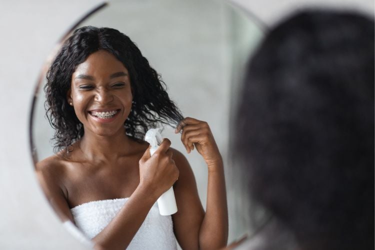 Reflexo de mulher de pele negra no espelho, usando toalha e aplicando spray de embalagem branca no cabelo