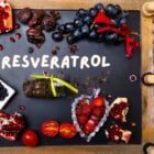 Foto de mesa com detalhes em madeira, contendo morango, uvas, mirtilo, e outras frutas roxas, além de rolhas de garrafas de vinho, bem como a palavra "resveratrol" em branco