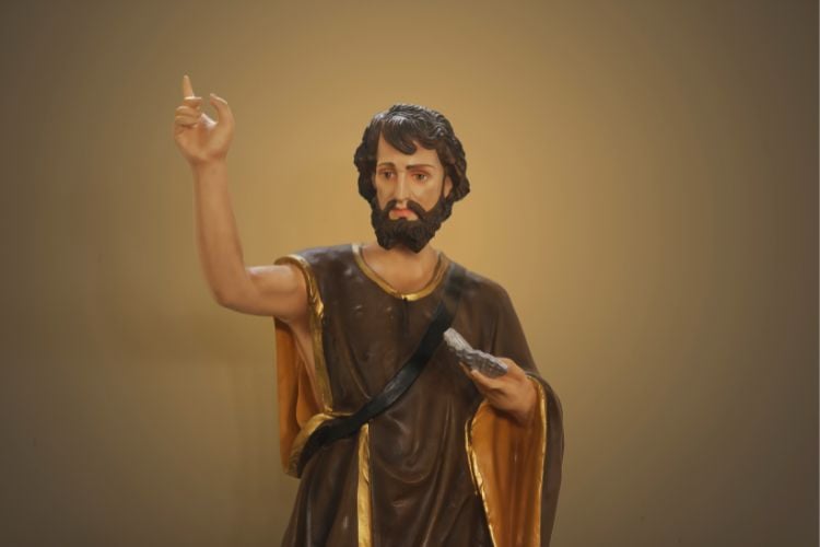 Foto de pequena escultura de São João, com manto marrom e um braço levantado