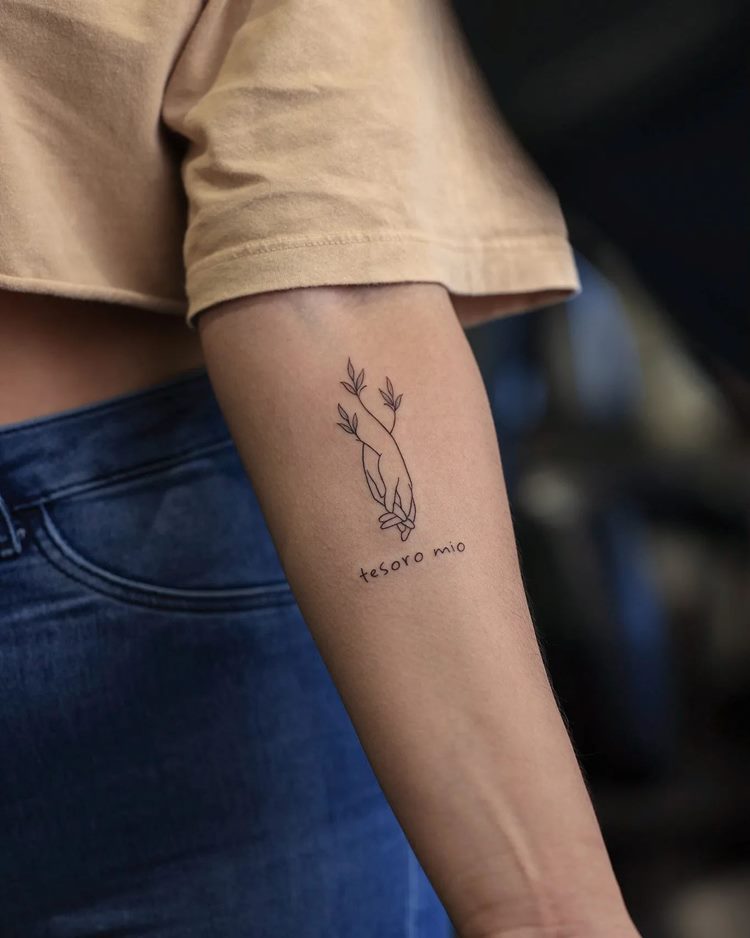Tatuagem de frase "tesouro meu" em espanhol com desenho de mãos de mãe e filho entrelaçando