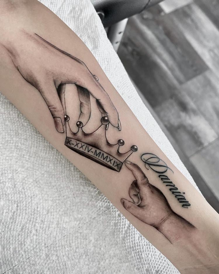 Tatuagem de mão com coroa e mão de bebê com nome de filha, cor preto