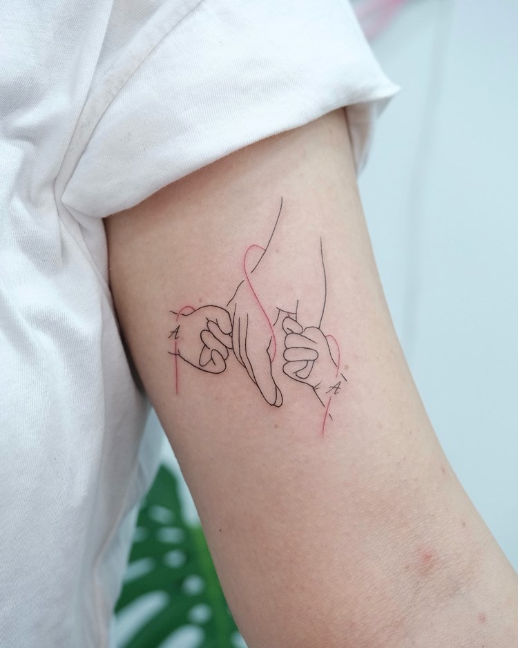 Tattoo de mãe em homenagem aos filhos: traços finos: mão segurando as mãos dos filhos em volta de um fio vermelho. Tattoo no braço