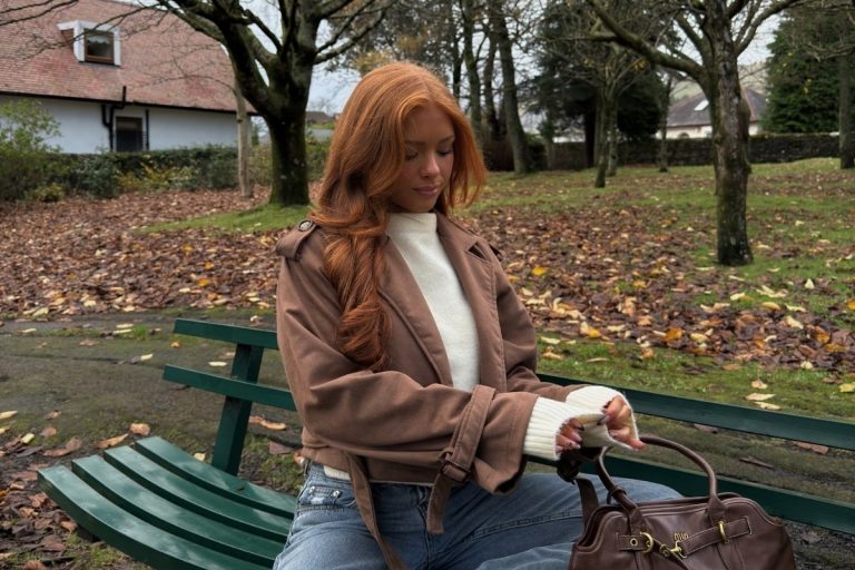 Mulher de cabelos ruivos longos sentada em banco verde em parque outonal. Usa jaqueta marrom, suéter branco e jeans. Segura bolsa marrom grande com detalhe dourado. Folhas caídas pelo chão e casa branca ao fundo.