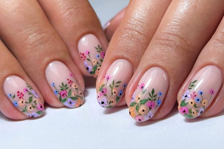 Unhas decoradas com esmalte nude, adornadas com delicadas flores coloridas, incluindo rosas, margaridas e folhas verdes, formando um design artístico e sofisticado. O fundo claro destaca os detalhes florais, criando um visual elegante e feminino para as mãos.