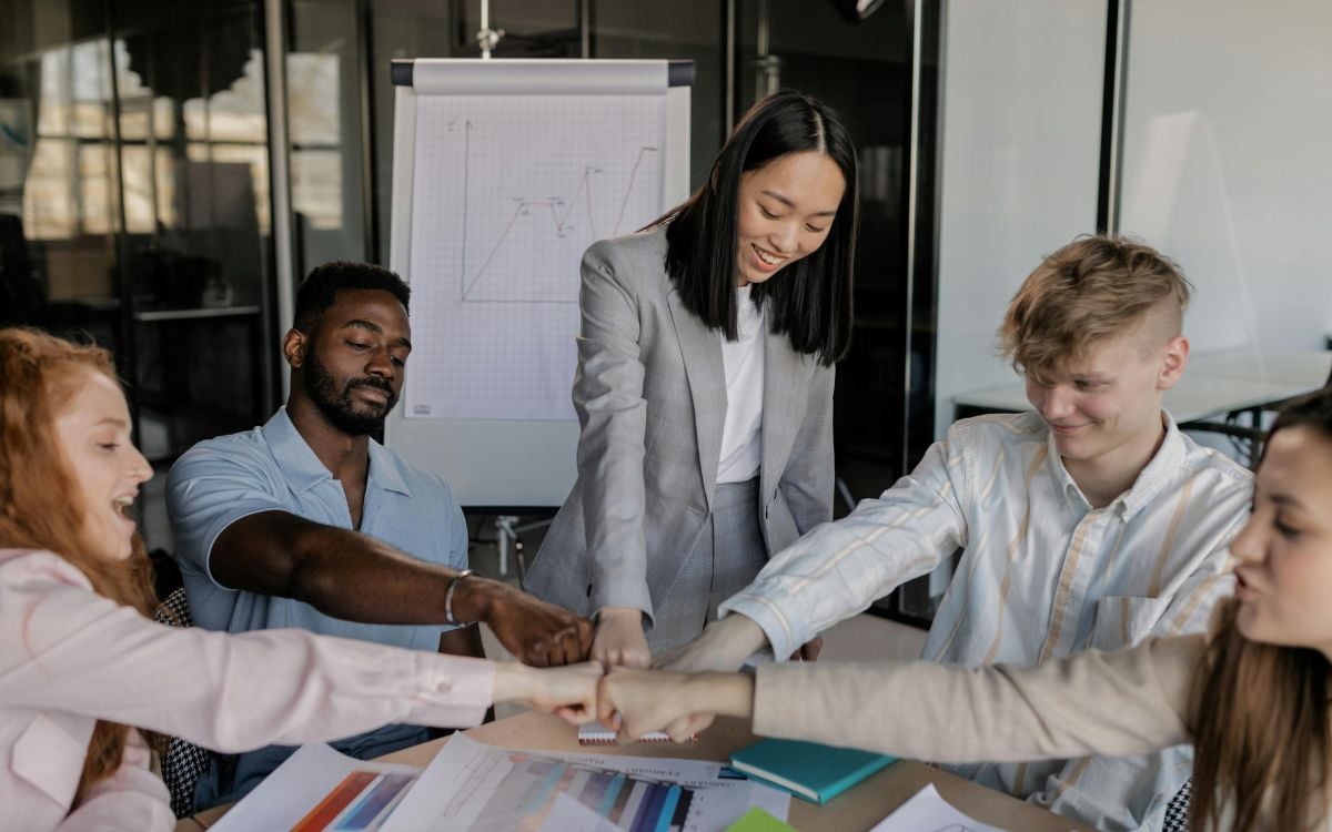 Foto de 5 profissionais em torno de mesa de reunião. Eles colocam as mãos uma em cima da outra, simbolizando espiritualidade nas empresas