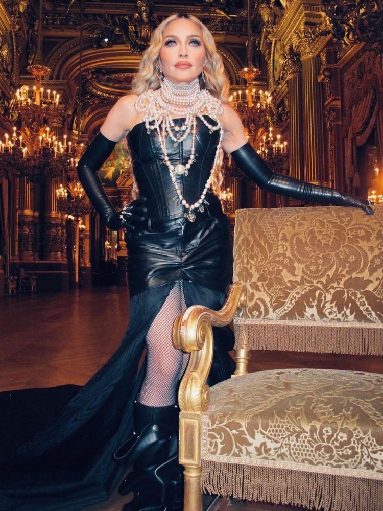 Foto de Madonna em palácio decorado com tons de dourado. A rainha do pop usa colar de pérolas, corset, saia e luvas de couro preto