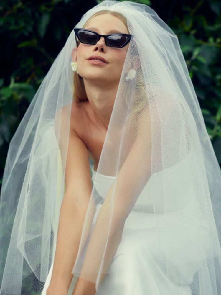 Mulher de pele clara usando óculos de sol, vestido de noiva sem mangas e véu