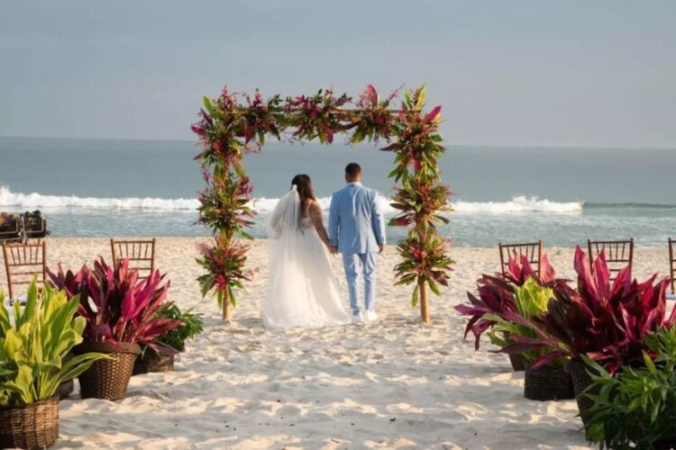 Imagem de noivo e noiva de costas para a foto e de frente para o mar, com céu limpo, flores coloridas e arco enfeitado com flores