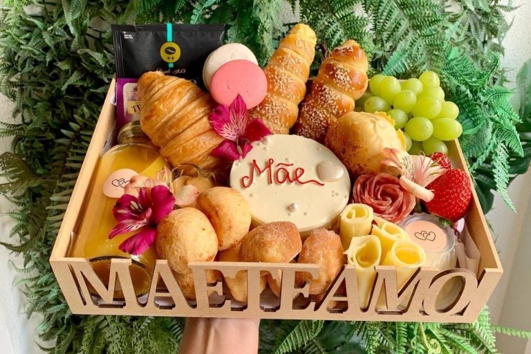 Uma cesta de café da manhã com pães, croissants, queijos, frutas, macarons e suco. No centro, uma bolacha decorada com "Mãe". A cesta tem flores ornamentais e o recorte "MÃE TE AMO" na lateral, sobre folhagens verdes.