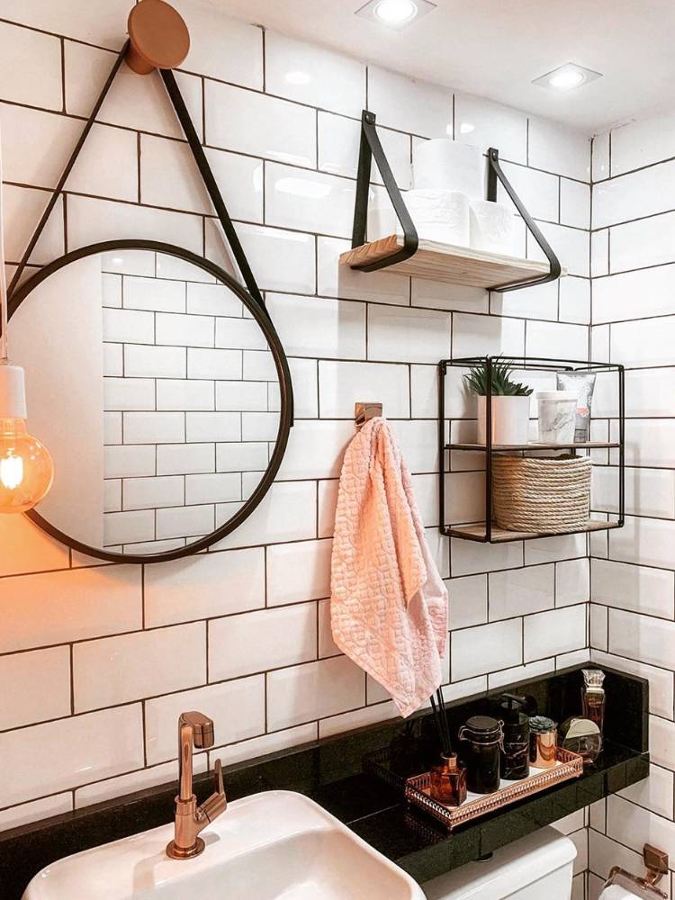 Banheiro de azulejos brancos com espelho redondo com alça de couro preta, prateleira branca com alça de couro preta, toalha, pia e decorações