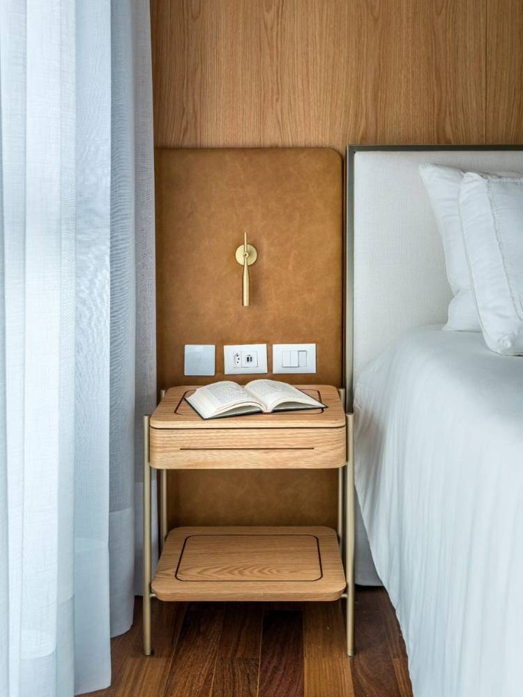 Cabeceira de couro marrom com luminária, cômoda de madeira, ao lado de cama com lençol branco