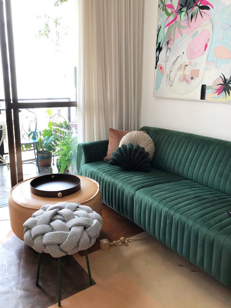 Sala com sofá verde de veludo, puff cinza, quadro colorido, almofadas e puff de centro em couro marrom
