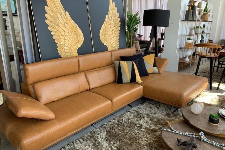 Sofá marrom de couro em frente à parede azul com asas douradas. O sofá conta com almofadas e está disposto próximo a tapete felpudo bege