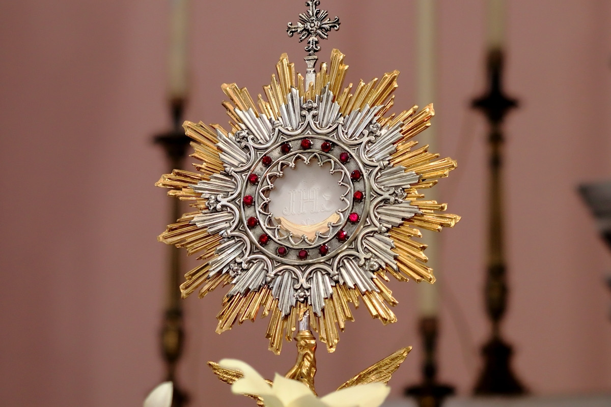 Imagem de um ostensório dourado com detalhes prateados e pedras vermelhas incrustadas em um círculo ao redor de uma hóstia no centro, simbolizando o corpo de Cristo. Ao fundo, velas desfocadas em um ambiente litúrgico.