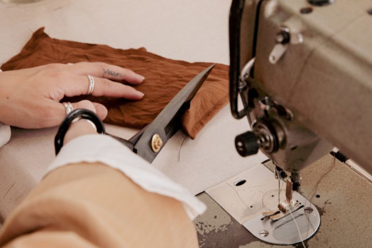 Foto de mão cortando tecido marrom ao lado de máquina de costura de escola de moda