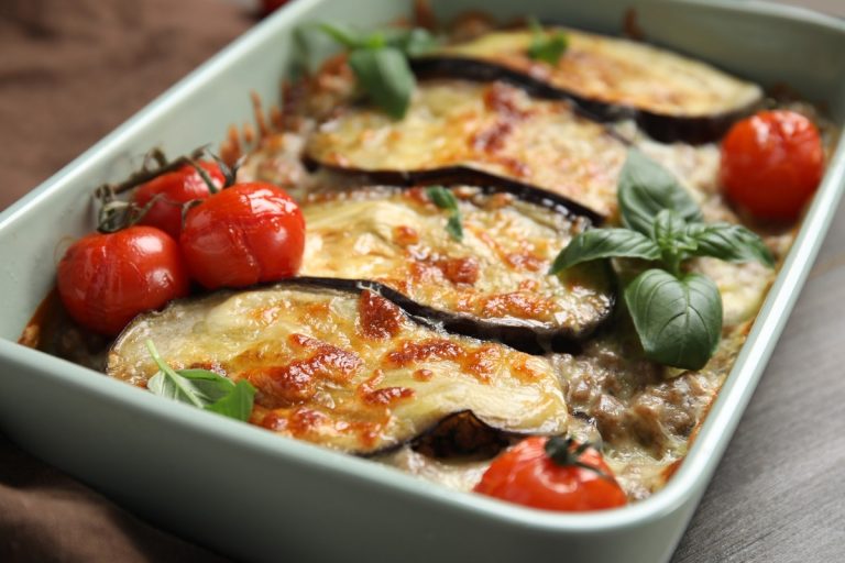 Uma travessa verde contendo uma berinjela assada com queijo derretido e gratinado. Decoração com tomates cereja vermelhos e folhas de manjericão fresco. O prato está em uma superfície de madeira, criando um aspecto apetitoso e convidativo.