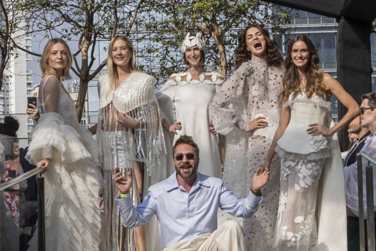 Mulheres de pele clara usando diferentes vestidos de noiva e sorrindo, enquanto o estilista Lucas Anderi está usando camisa azul e terno bege, abaixado em frente às modelos