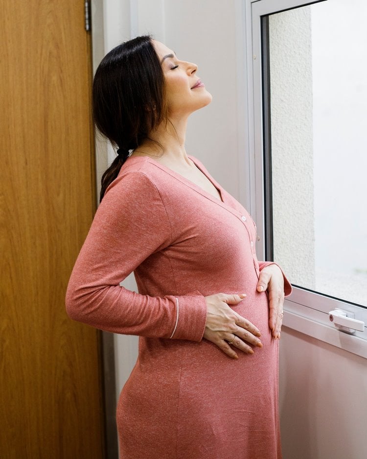 Apresentadora Nadja Haddad grávida de gêmeos com as mãos na barriga, usando camisola rosa, de frente à janela em hospital Pro Matre