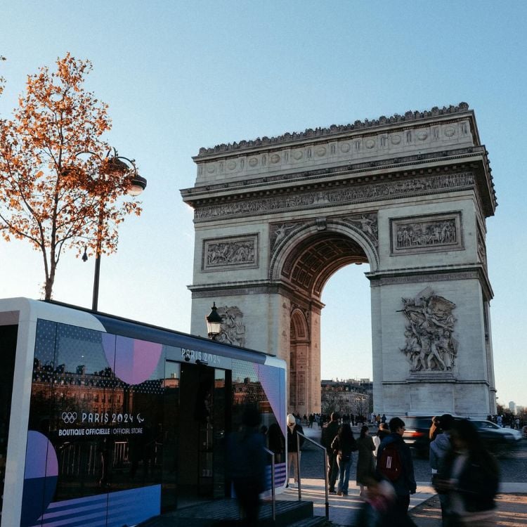 Foto de um dia com céu claro e limpo, Arco do Triunfo ao fundo, pessoas e ônibus escrito "Paris 2024"