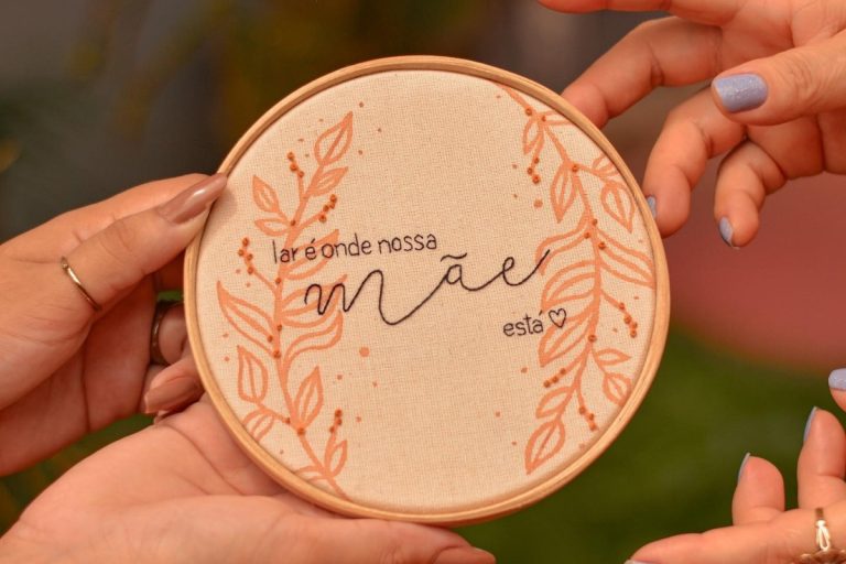 Mãos de pele clara segurando bastidor bordado com frase "Lar é onde nossa mãe está"