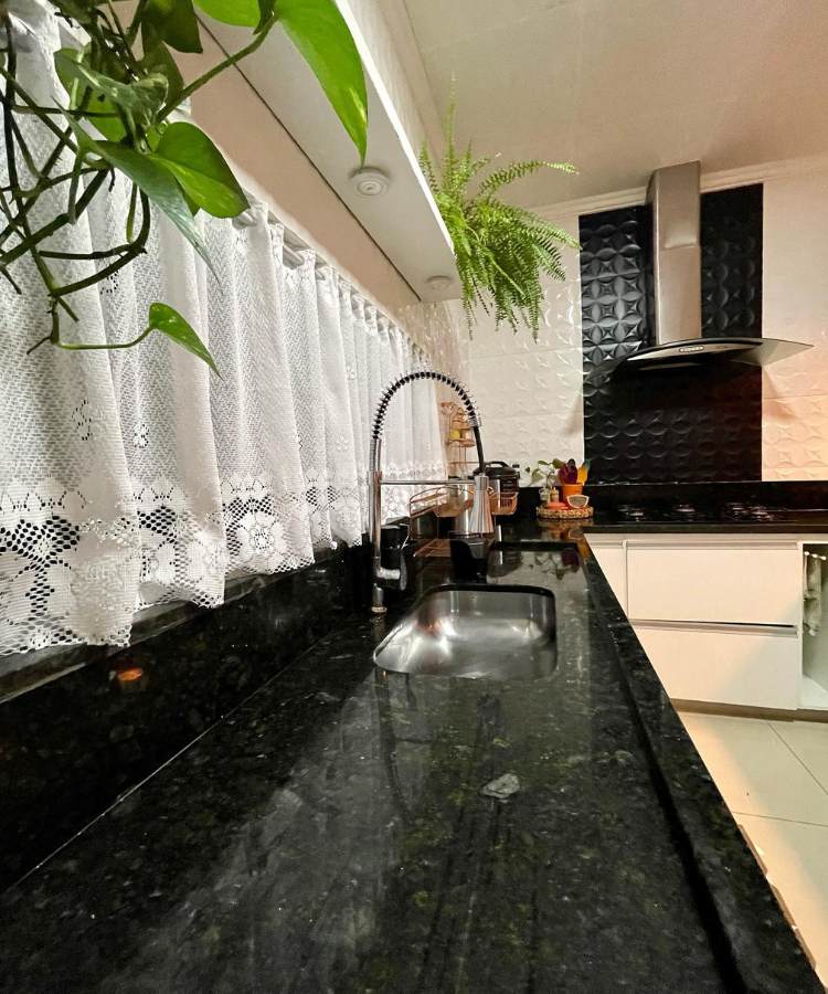 Cozinha com pia de pedra preta, cortina de renda e plantas jiboia e samambaia penduradas