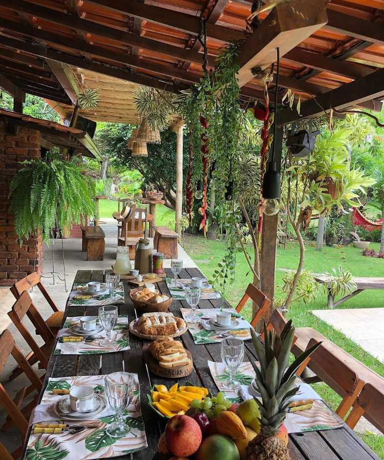 Mesa com café da manhã em uma casa rural com diversas plantas penduradas, inclusive, samambaia