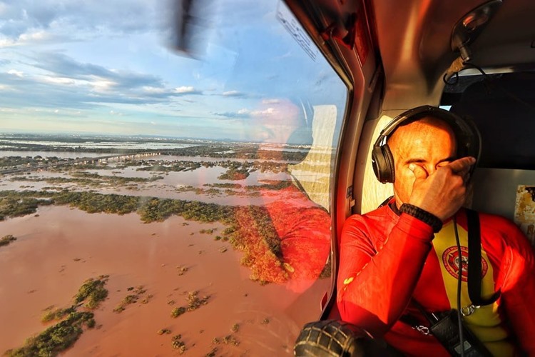 A foto mostra um bombeiro, dentro de um helicóptero. Ele esfrega os olhos, em sinal de aflição ou cansaço. À esquerda, pela janela, vemos uma grande área alagada.