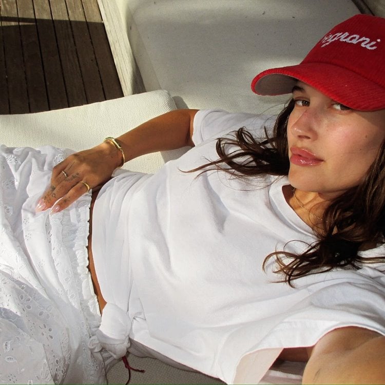 Hailey Bieber deitada com roupa branca e boné vermelho