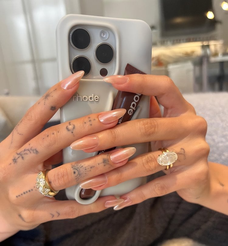 Mãos de Hailey Bieber segurando Iphone com capinha que segura gloss, ela usa unhas gladez donut