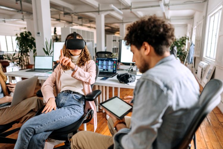 Foto de mulher usando óculos de realidade virtual ao lado de designer de games segurando tablet e fazendo teste