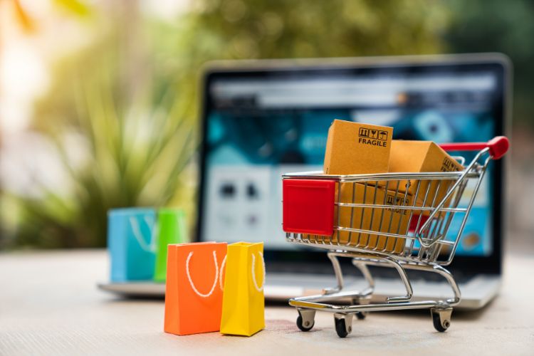 Foto de notebook aberto em e-commerce, rodeado de carrinho de compras, caixas de encomendas e sacolas coloridas