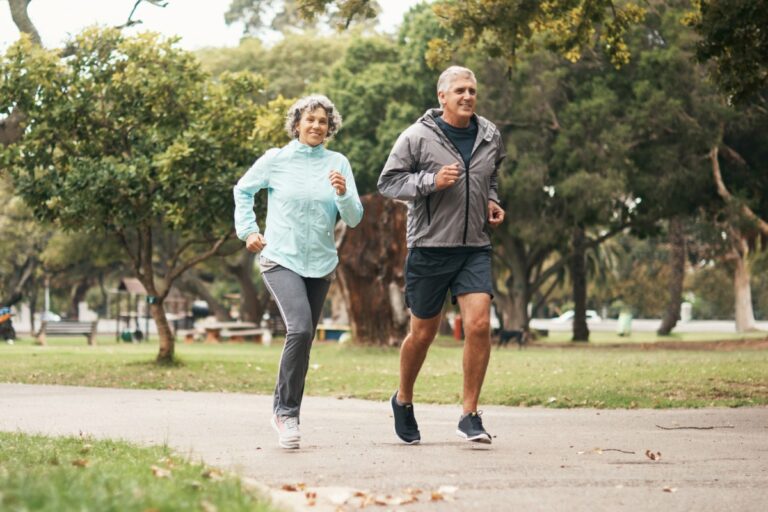 Mulher e homem idosos (ela de blusa azul e calça preta e ele de blusa cinza e bermuda preta) correndo em um parque