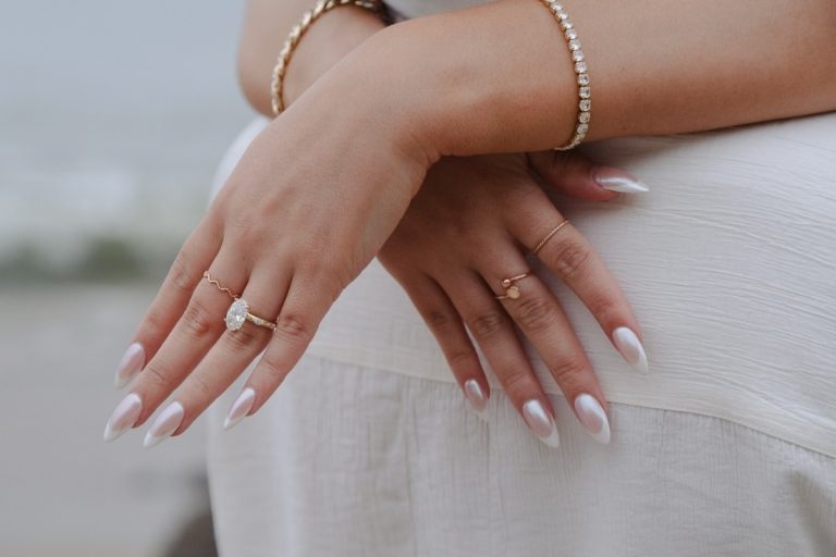 Mãos femininas com unhas em estilo amendoado e esmalte perolado. Anéis dourados delicados e uma pulseira de pedras brilham, destacando a sofisticação. O fundo desfocado sugere um ambiente ao ar livre, criando um cenário romântico e elegante.