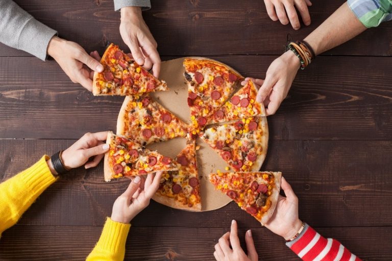 Mãos pegam pedaços de pizza que está em uma mesa de madeira.