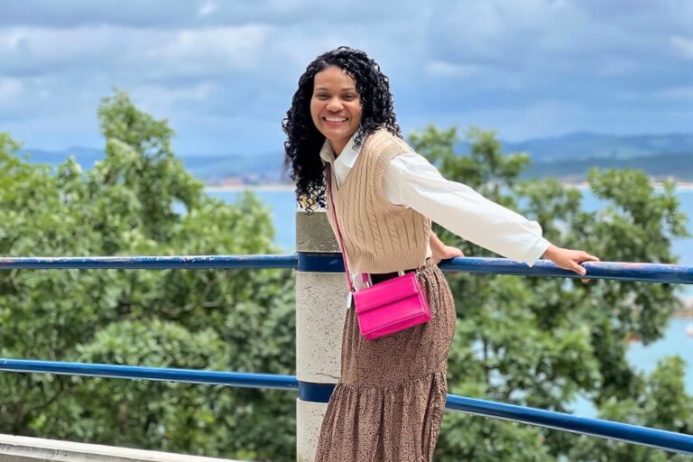 A pessoa na foto veste roupas que se enquadram no estilo da moda evangélica. Ela usa uma blusa branca de mangas compridas, um colete bege e uma saia marrom com detalhes plissados. Uma bolsa rosa choque completa o visual. O cenário ao fundo mostra um céu parcialmente nublado e um corpo de água.