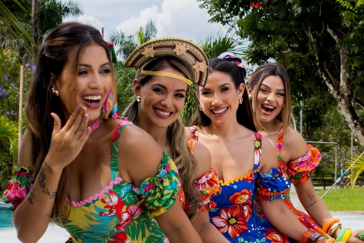 Quatro mulheres sorridentes em roupas festivas e coloridas posam ao ar livre, cercadas por vegetação tropical. Elas usam vestidos com estampas florais vibrantes e acessórios como laços e chapéus. O ambiente transmite alegria e celebração em um cenário tropical iluminado.
