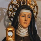 A imagem mostra Santa Clara de Assis com um hábito marrom e véu preto. Ela segura um ostensório dourado com uma hóstia branca. Santa Clara tem um rosto sereno e usa uma auréola dourada decorada com folhas. O fundo é neutro, destacando a figura da santa.
