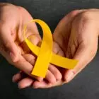 A imagem mostra duas mãos segurando uma fita amarela em forma de laço. As mãos estão em um fundo escuro, e a fita amarela é destacada no centro da imagem. O laço amarelo é frequentemente associado a campanhas de conscientização sobre saúde mental e prevenção do suicídio.
