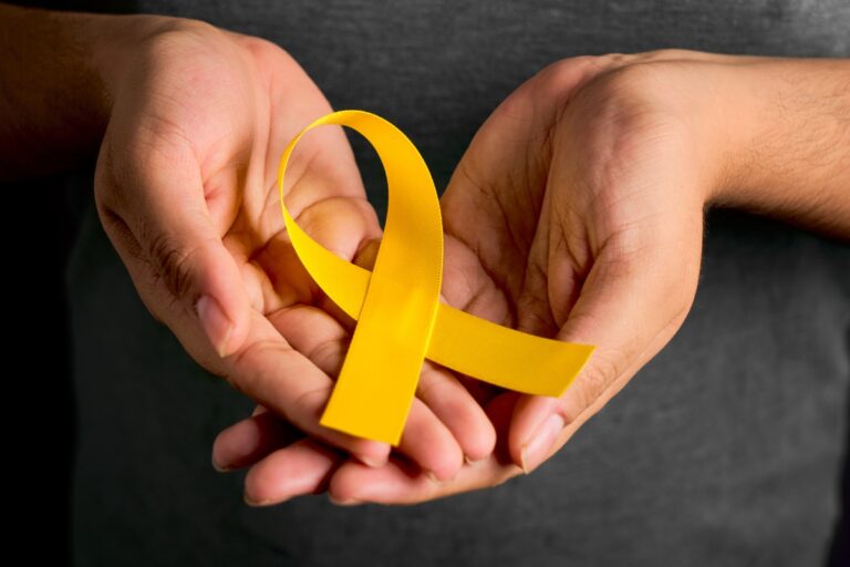 A imagem mostra duas mãos segurando uma fita amarela em forma de laço. As mãos estão em um fundo escuro, e a fita amarela é destacada no centro da imagem. O laço amarelo é frequentemente associado a campanhas de conscientização sobre saúde mental e prevenção do suicídio.