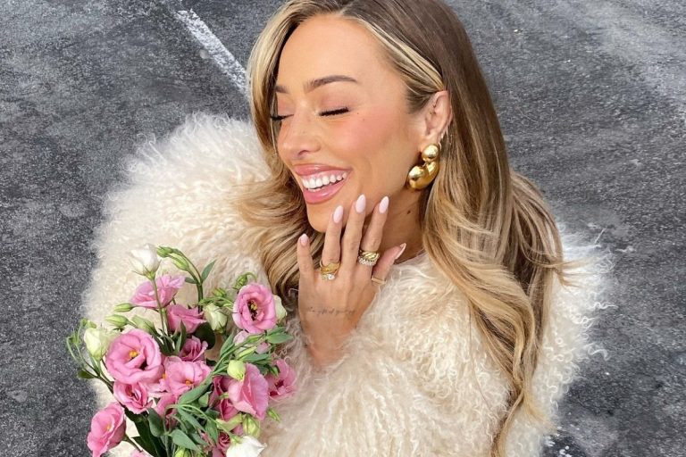 Mulher sorridente com cabelos loiros, usando brincos dourados, anéis e blusa felpuda branca. Ela segura um buquê de flores rosa e brancas, expressando alegria em um ambiente externo com chão cinza ao fundo.