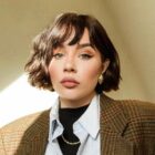 Uma pessoa de cabelo curto e escuro, com maquiagem destacando olhos e lábios, veste blazer xadrez marrom sobre camisa listrada azul e gola preta. Usa brincos dourados e colar. A luz suave ilumina o rosto, criando uma atmosfera elegante.