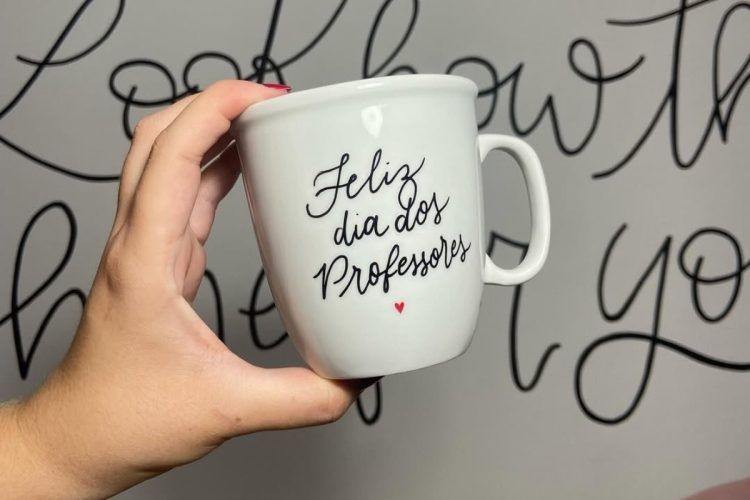 Pessoa de pele clara segurando caneca escrito "Feliz Dia Dos Professores"