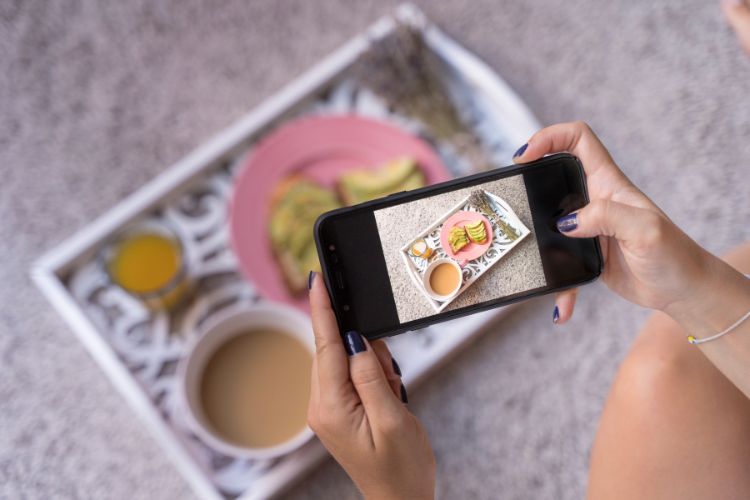 A imagem mostra mãos segurando um smartphone, fotografando uma bandeja de café da manhã. Na bandeja, há uma xícara de café, suco de laranja, abacate e biscoitos em um prato rosa. O ambiente tem um tom acolhedor e relaxante.