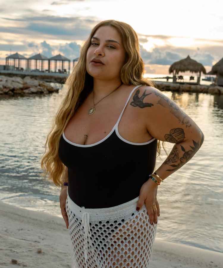 A imagem mostra uma mulher em uma praia, ao pôr do sol. Ela veste um maiô preto e uma saia de crochê branca. Seu cabelo é longo e ondulado, e ela tem tatuagens nos braços. O cenário inclui palhoças e um céu nublado.Influenciadoras plus size