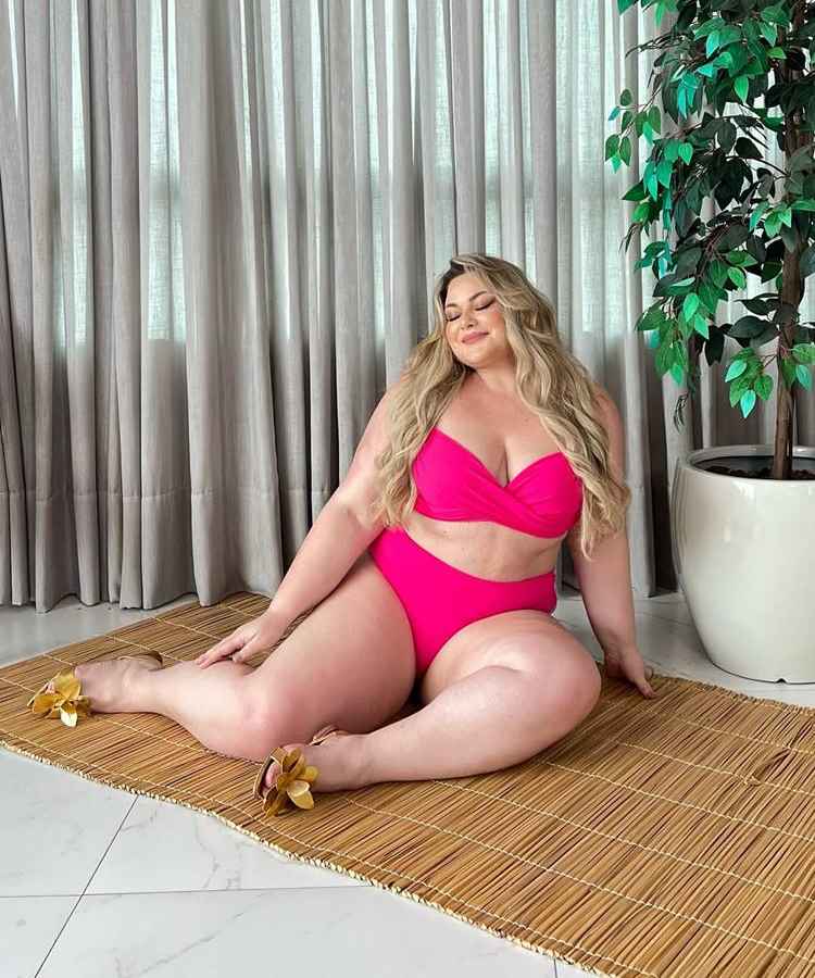 A imagem mostra uma mulher sentada em um tapete de palha, usando um biquíni rosa vibrante. Ela está sorrindo, com longos cabelos loiros soltos. Ao lado, uma planta em vaso complementa o ambiente interno, que tem cortinas cinza ao fundo.Influenciadoras plus size