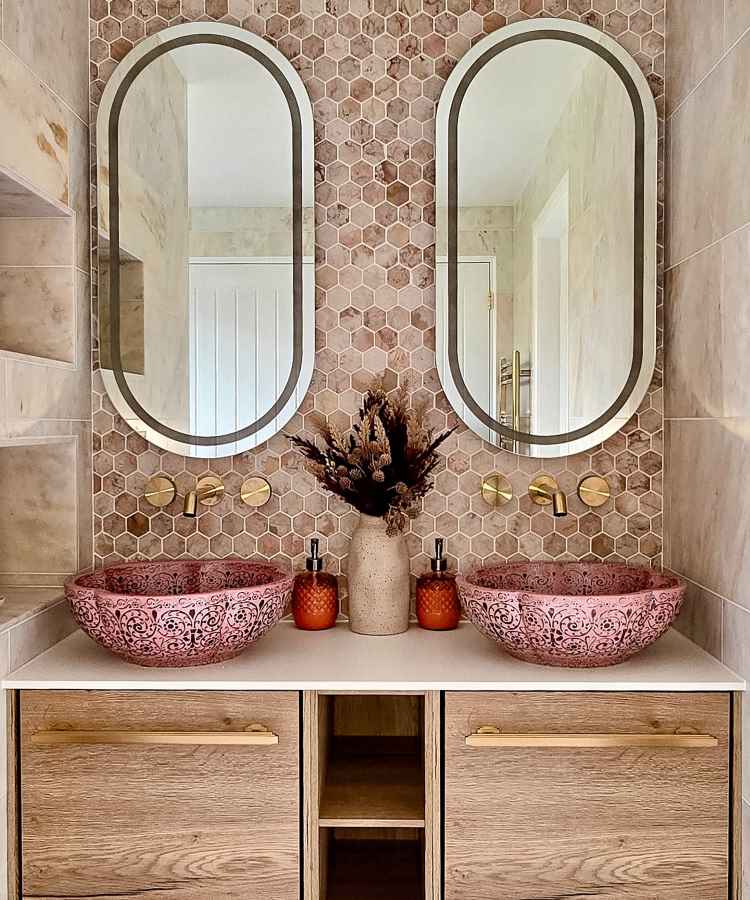 Banheiro sofisticado com pia dupla de louça rosa ornamentada, diante de espelhos ovais. A parede é revestida com azulejos hexagonais em tons terrosos de Mocha Mousse na decoração. Abaixo, armários de madeira clara oferecem armazenamento. Detalhes dourados nos acessórios e torneiras complementam o design elegante e acolhedor.